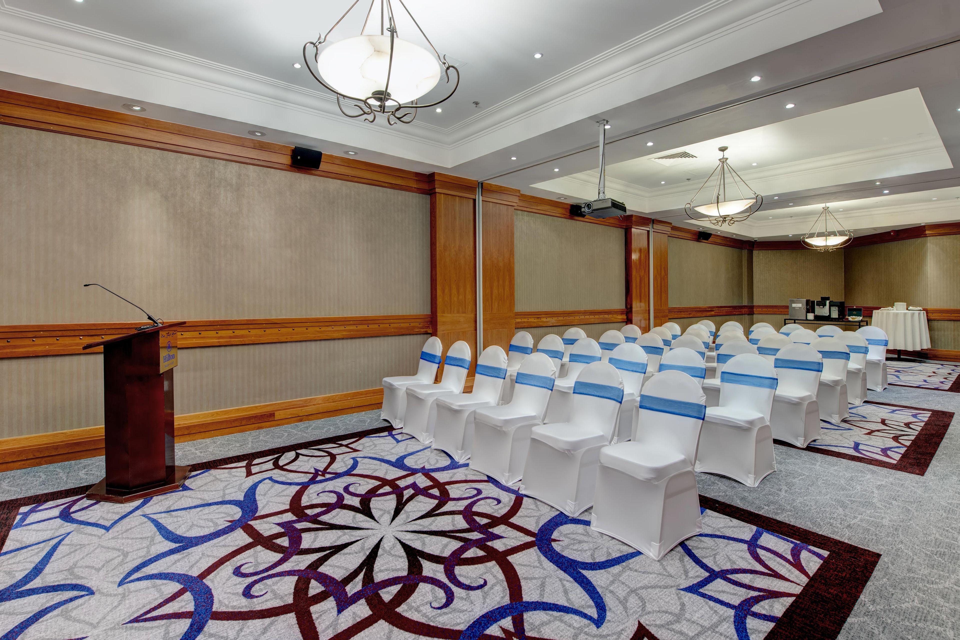 Une salle de conférence avec des rangées de chaises blanches, un pupitre et un tapis orné d’un motif géométrique.