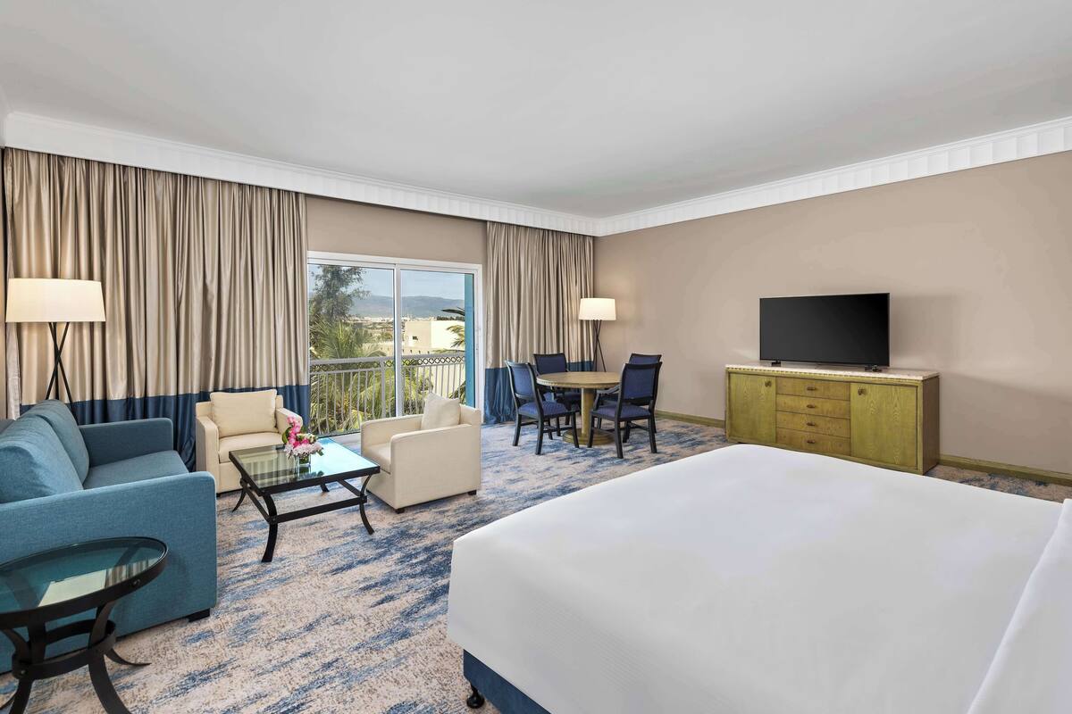 Habitación de hotel con una cama grande, un sofá, una mesita, un televisor en un mueble de madera y un balcón con cortinas.