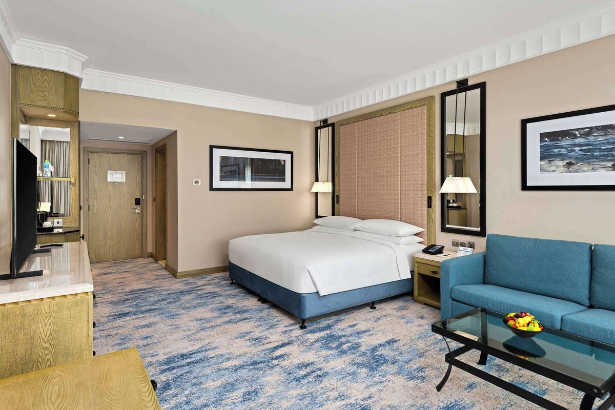 Habitación de hotel con cama, sofá, mesa de centro de vidrio con fruta, televisor y escritorio con lámpara.