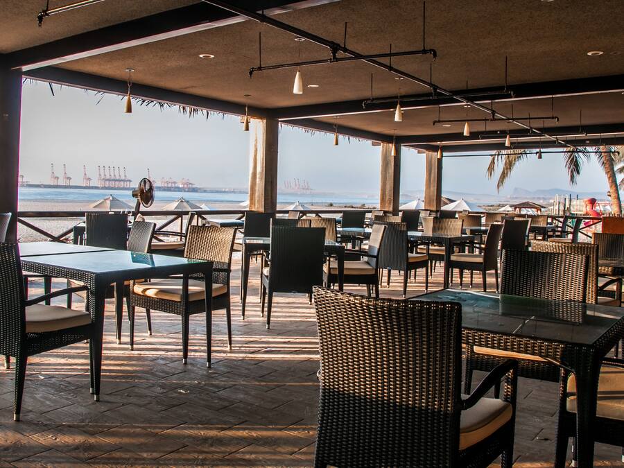 An outdoor seating area with tables and chairs, overlooking a body of water with industrial structures in the distance.