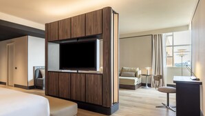A modern hotel room with a large bed, a TV mounted on a wooden panel wall, a seating area with a small table and chair, and a view of the city through large windows.