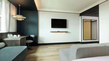 A modern hotel room with a wooden desk, a flat-screen TV mounted on the wall, a sofa, and a bed with striped bedding.