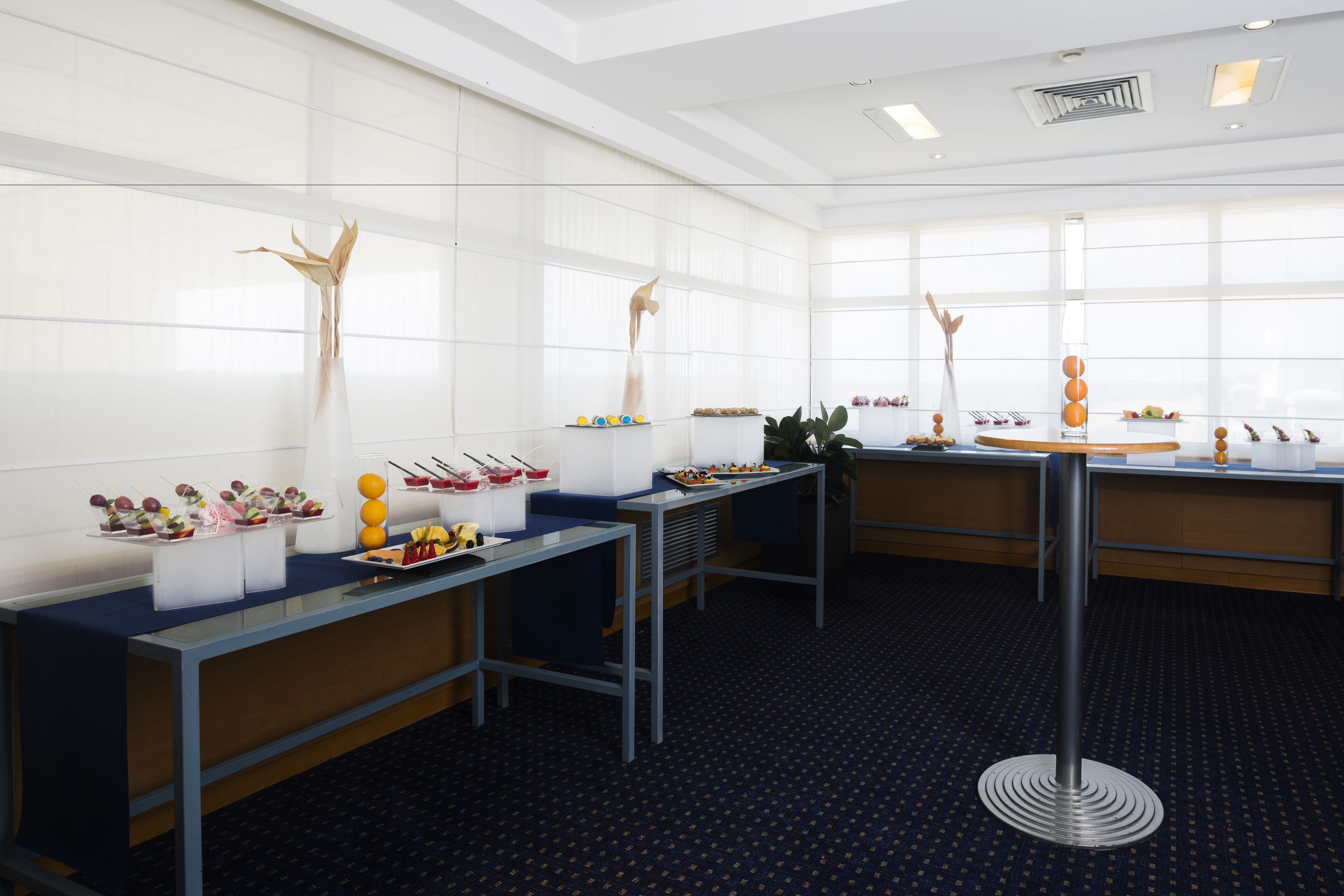 A modern hotel room with a large window, a round table with a unique base, and a buffet setup with various food items.
