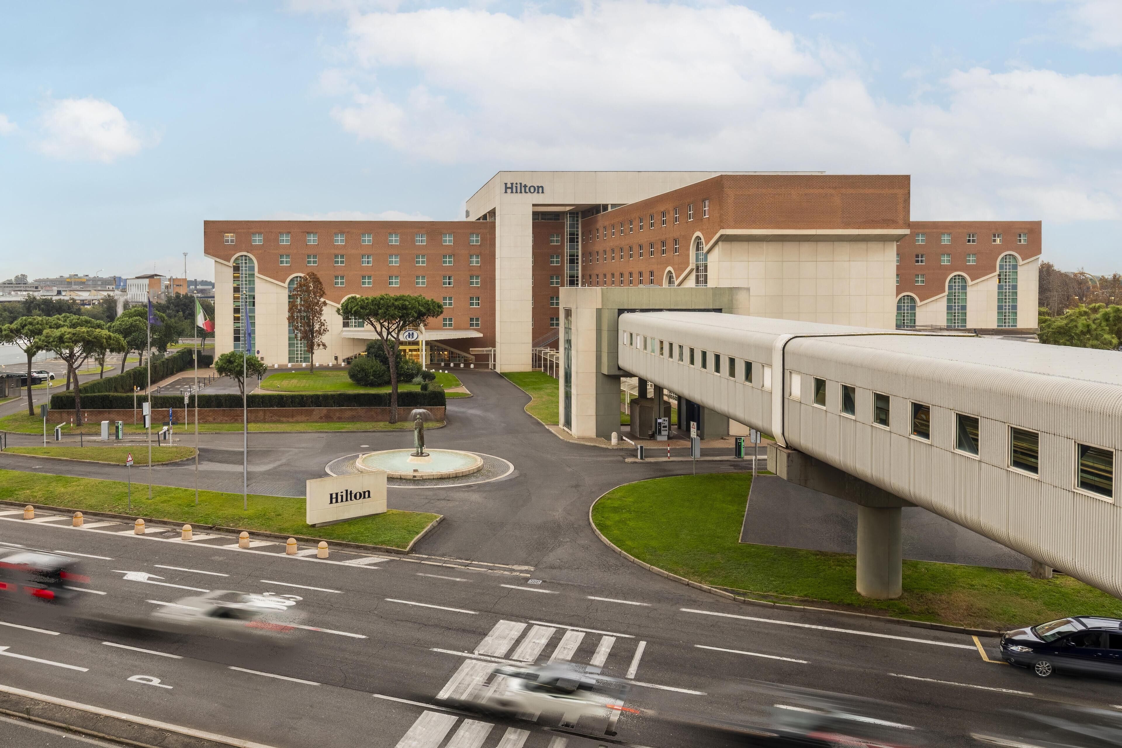 Un hôtel moderne, reconnaissable à son grand panneau Hilton, doté d’une cour intérieure et d’un pont piétonnier enjambant une route.