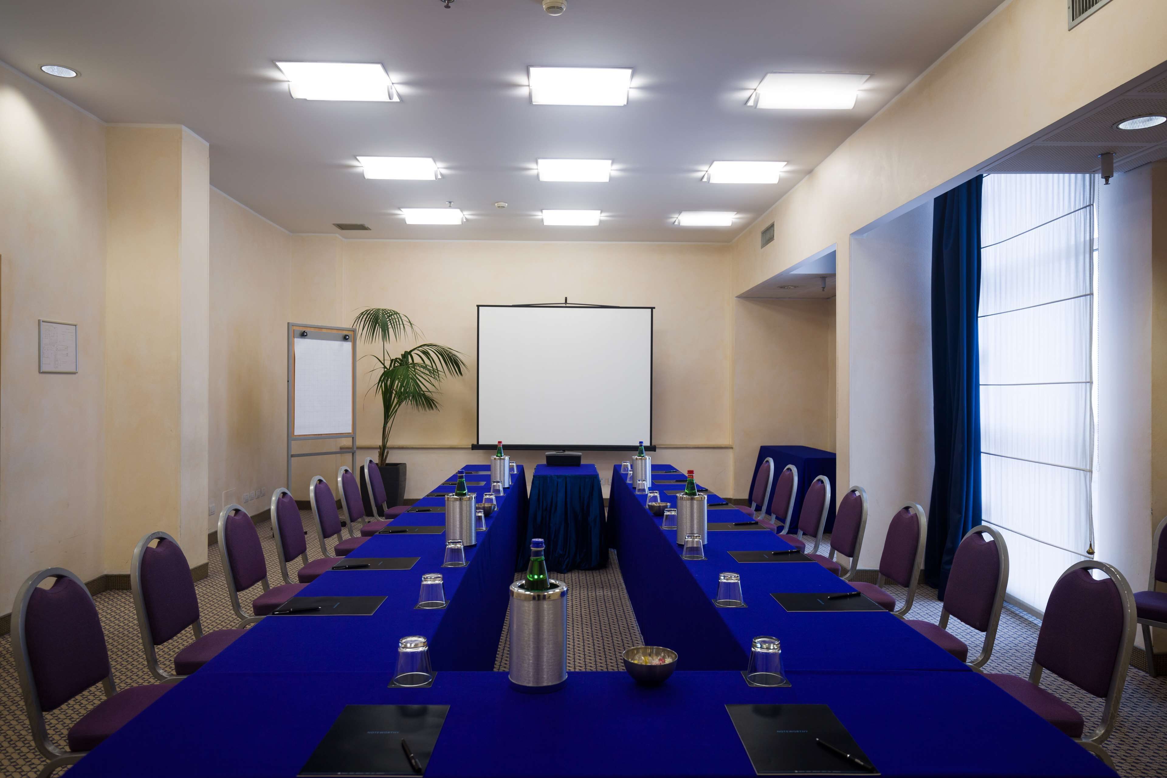 A conference room with a long table, chairs, a projector screen, and a plant.