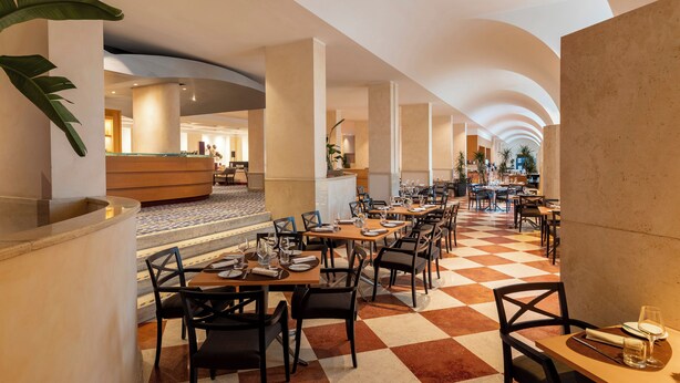 A spacious hotel lobby with a checkered floor, multiple dining tables, and a central reception area.