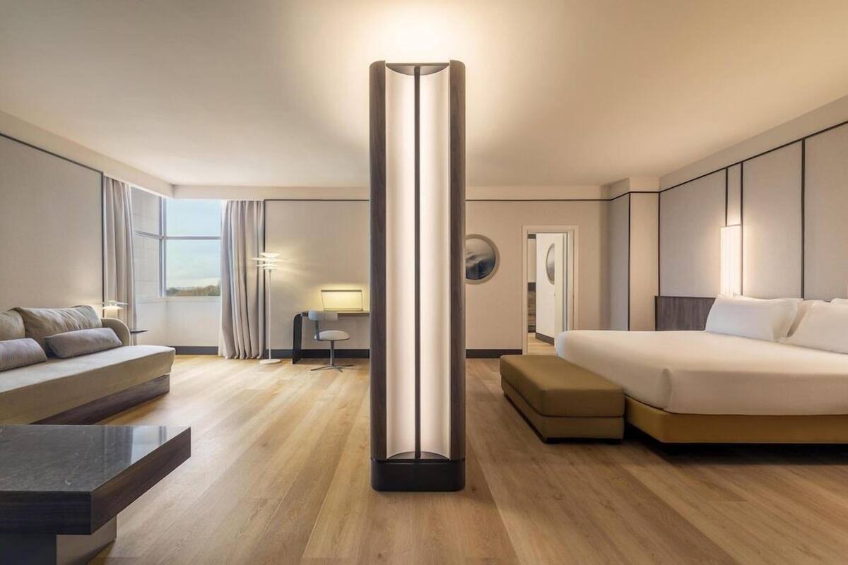 A modern hotel room with a central floor lamp, a sofa, a bed, and a desk with a chair.