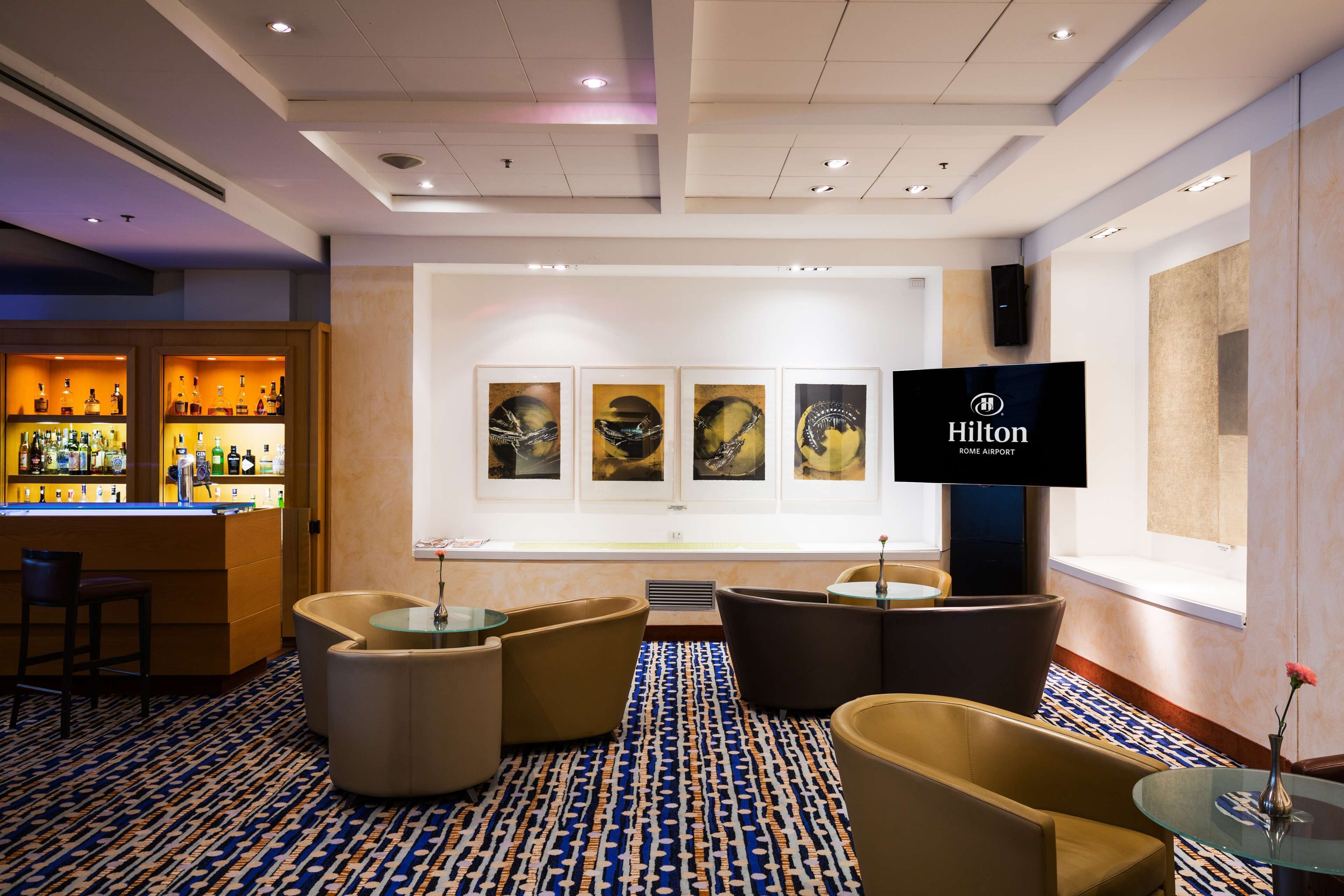 A modern hotel lobby with a bar area, comfortable seating, artwork on the wall, and a television displaying the hotel name.