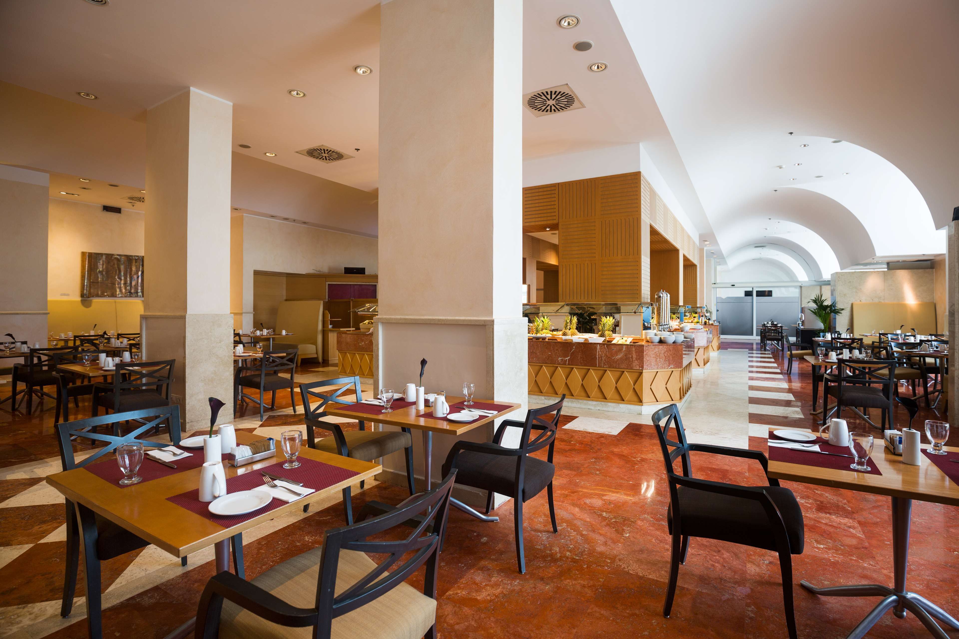 Un amplio comedor de hotel con varias mesas y sillas, una barra central y un techo alto con iluminación empotrada.