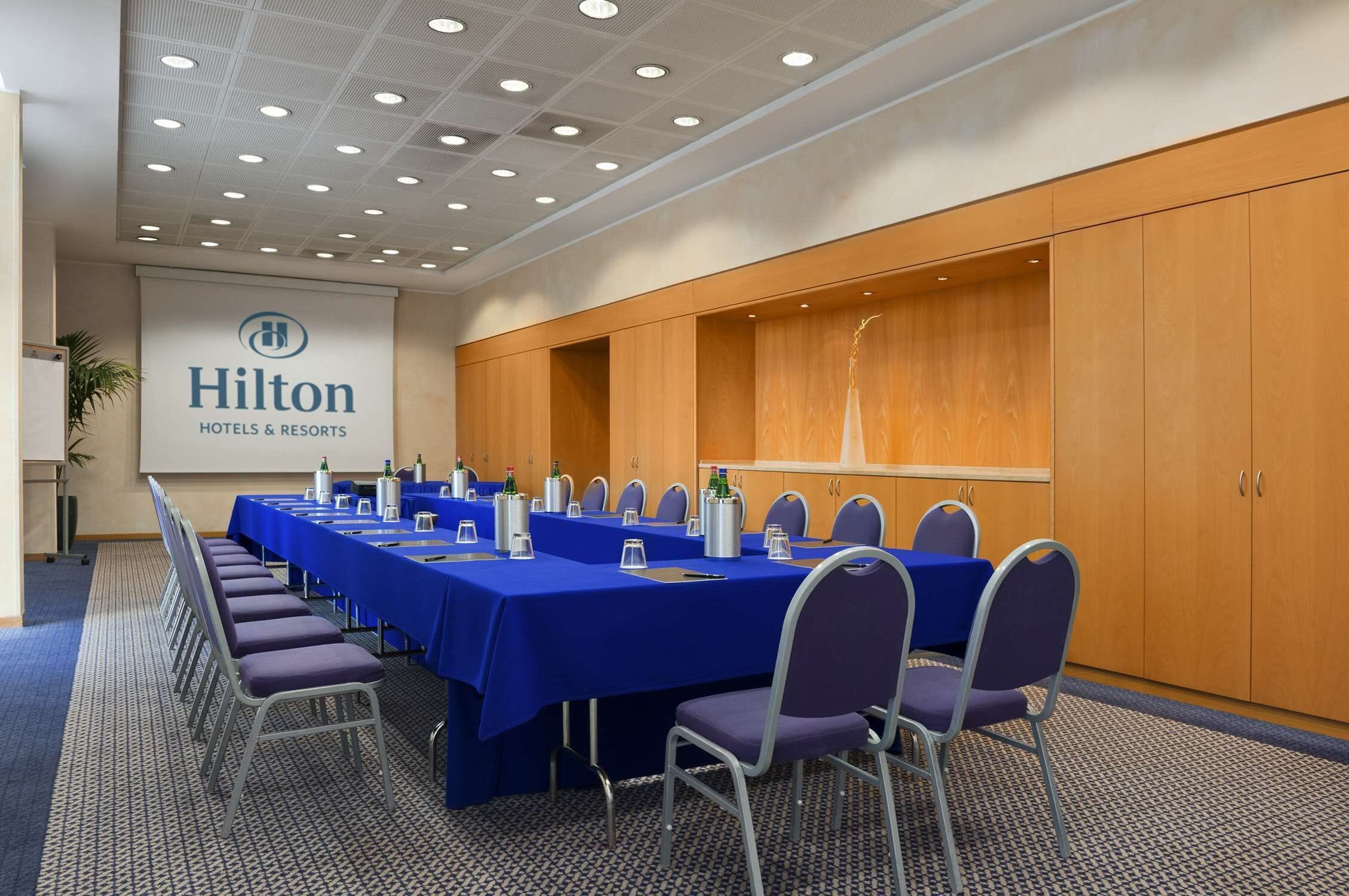 Un salón de conferencias con mantel azul, sillas dispuestas y un cartel de Hilton Hotels & Resorts.