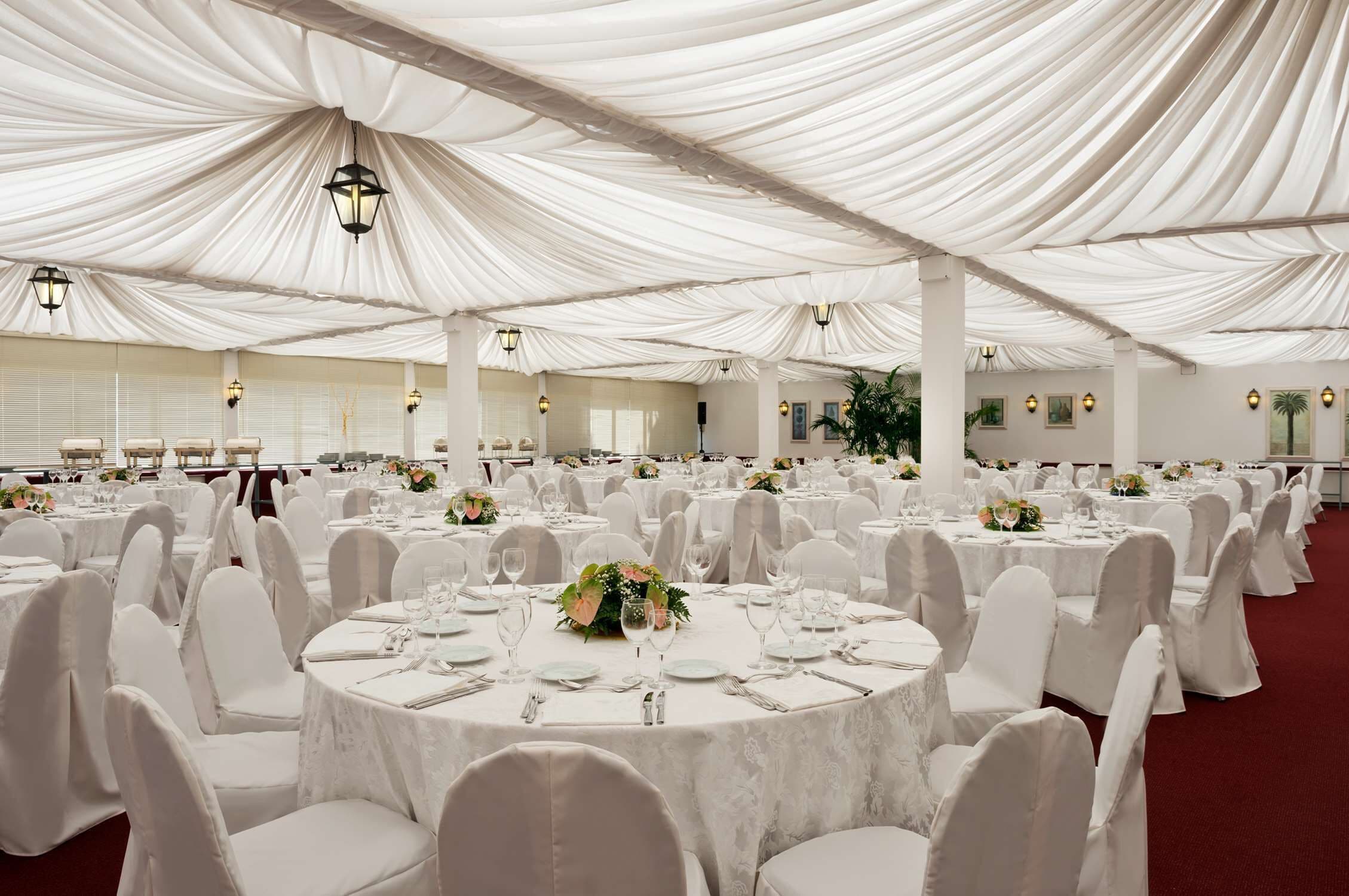 Un salón de banquetes con mesas redondas dispuestas para el servicio de comidas, techos cubiertos con tela blanca e iluminación fijada a la pared.