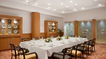 Un salón de conferencias con una larga mesa dispuesta para una reunión, rodeado de sillas, con platos decorativos en estanterías y un diseño de techo moderno.