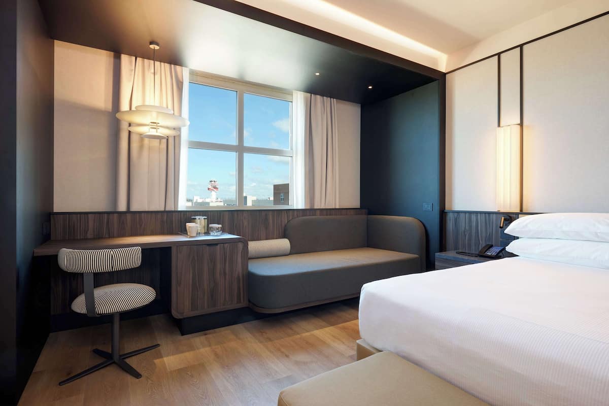 A modern hotel room with a bed, a sofa, a desk with a chair, and a window with curtains.