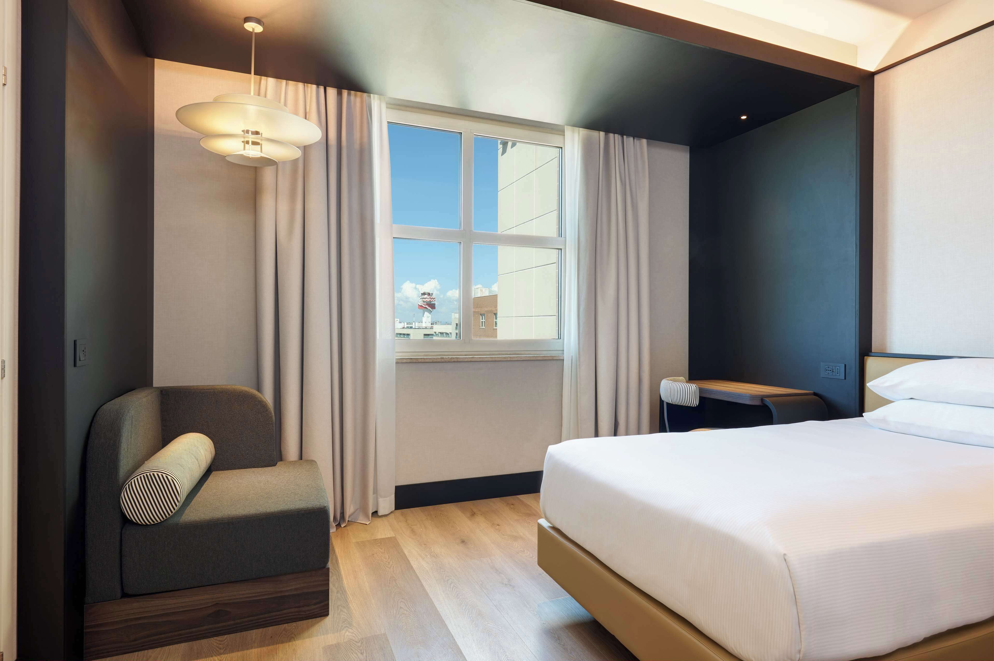A modern hotel room with a bed, a chair, a desk, and a window with curtains.