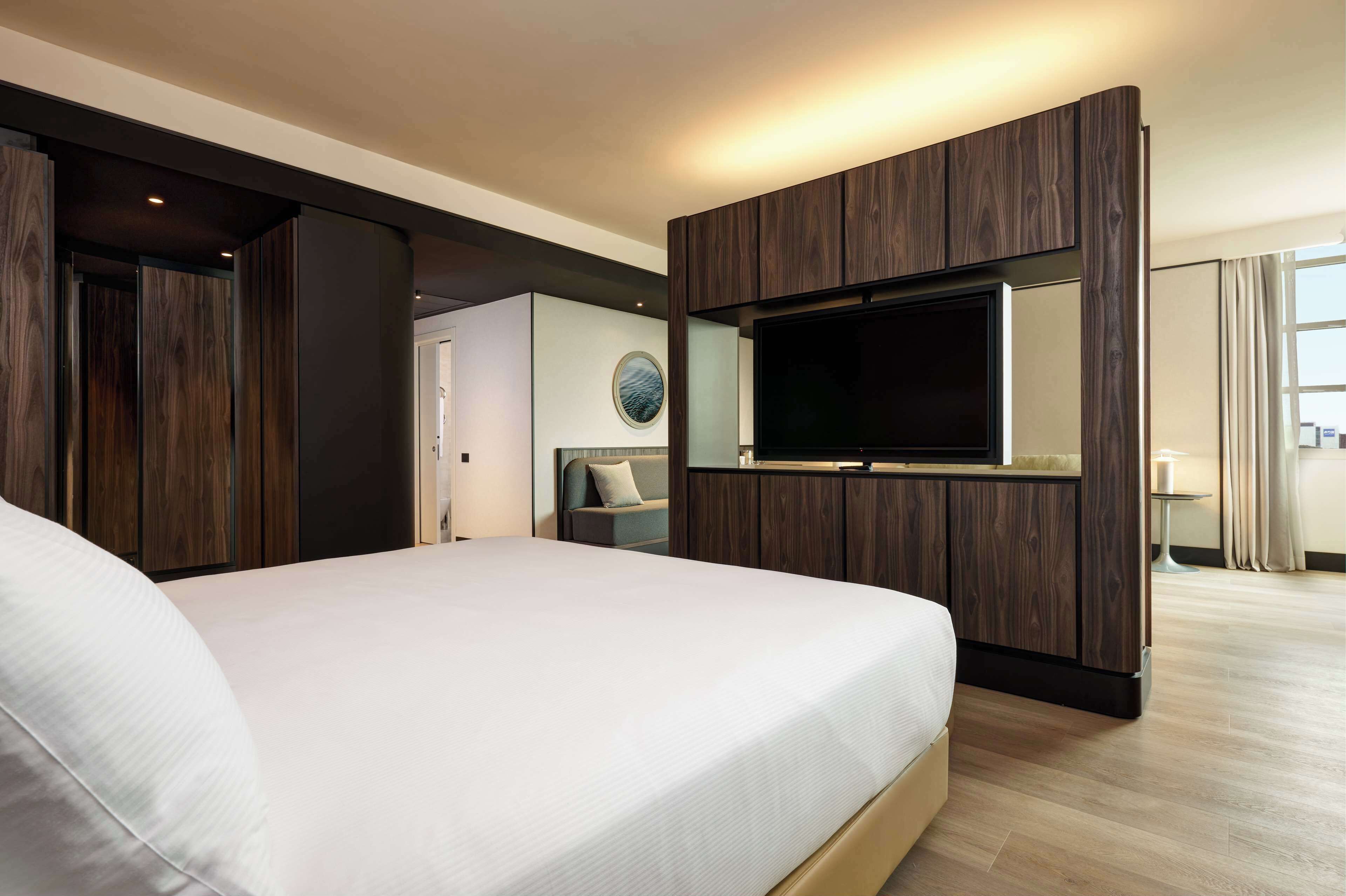 Una habitación de hotel moderna con una cama grande, un televisor de pantalla plana montado sobre un panel de madera, un pequeño rincón de descanso con un sof á y vistas a la ciudad desde la ventana.