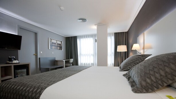Ein Hotelzimmer mit einem großen Bett, einem Schreibtisch, einem Flachbildfernseher, einer Kaffeemaschine und einem Fenster mit Vorhängen.