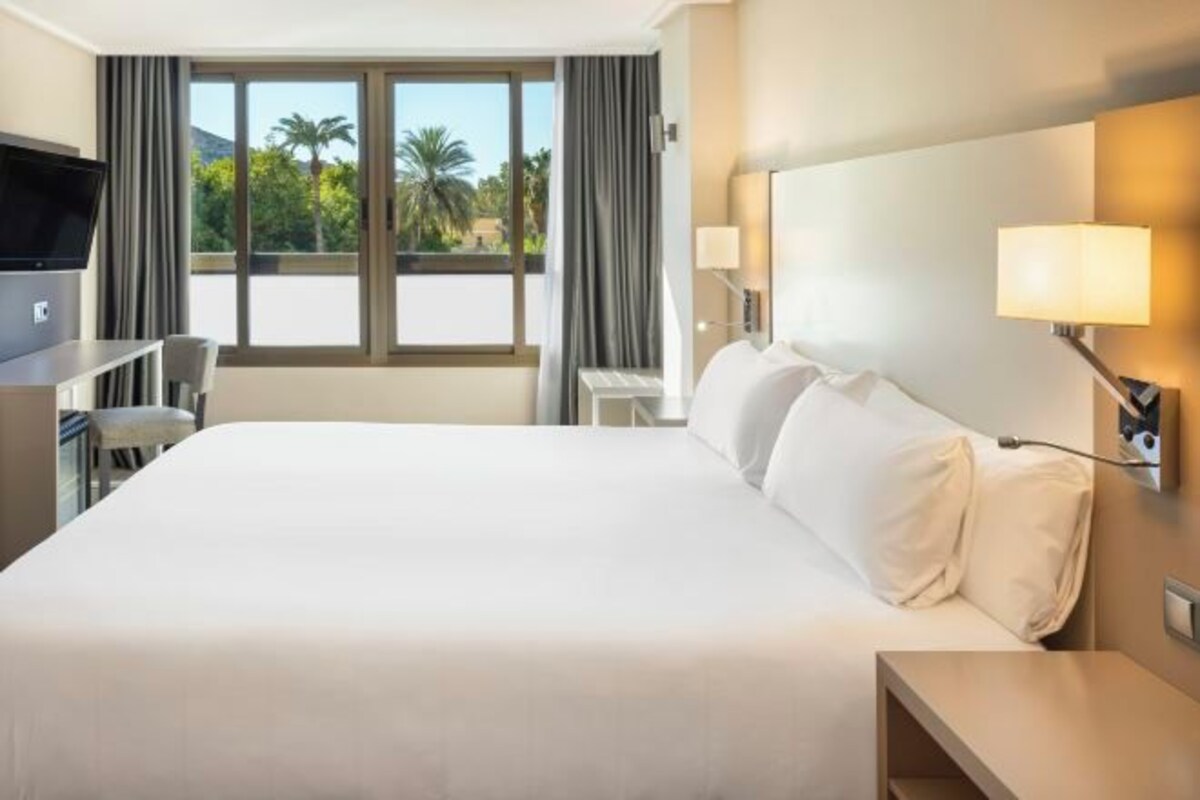 Une chambre d’hôtel avec un grand lit, un bureau avec une chaise, une télévision et une fenêtre avec des rideaux.
