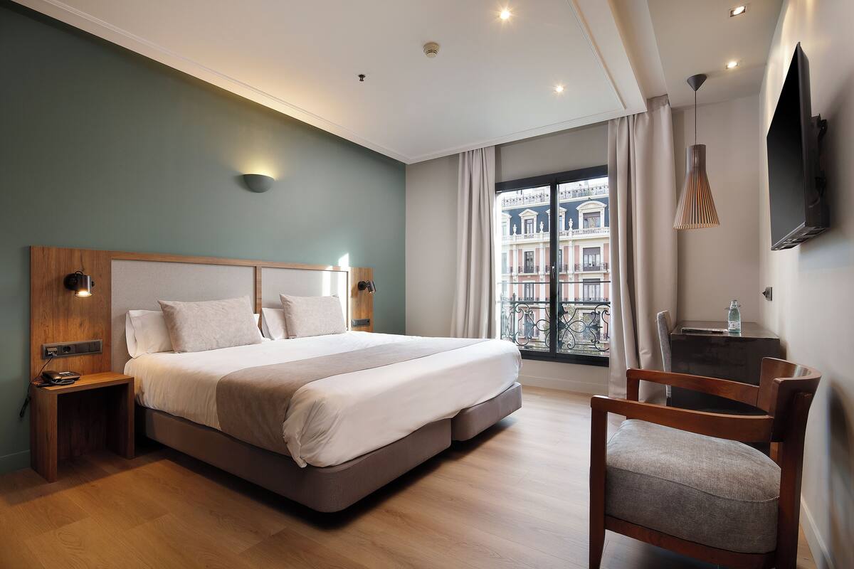 Une chambre d’hôtel moderne avec un grand lit, une table de chevet en bois, un bureau, une chaise et un téléviseur fixé au mur.