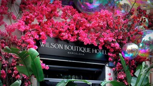 Exterior do Wilson Boutique Hotel em Barcelona, com fachada preta e placa, rodeado por flores rosa vibrantes e folhagem verde.