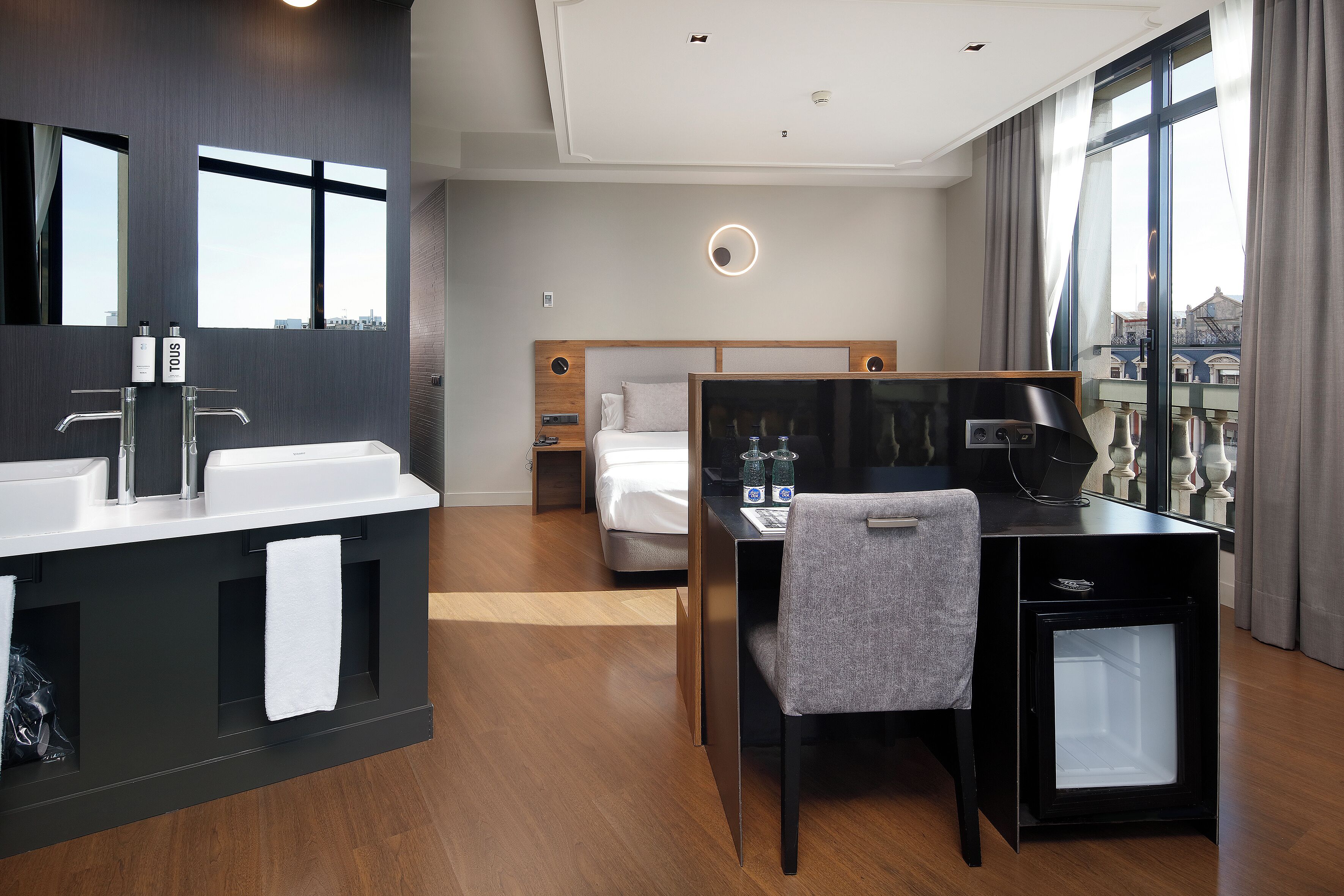 A modern hotel room with a large window, a desk with a chair, a bed, and a bathroom with a vanity and mirror.