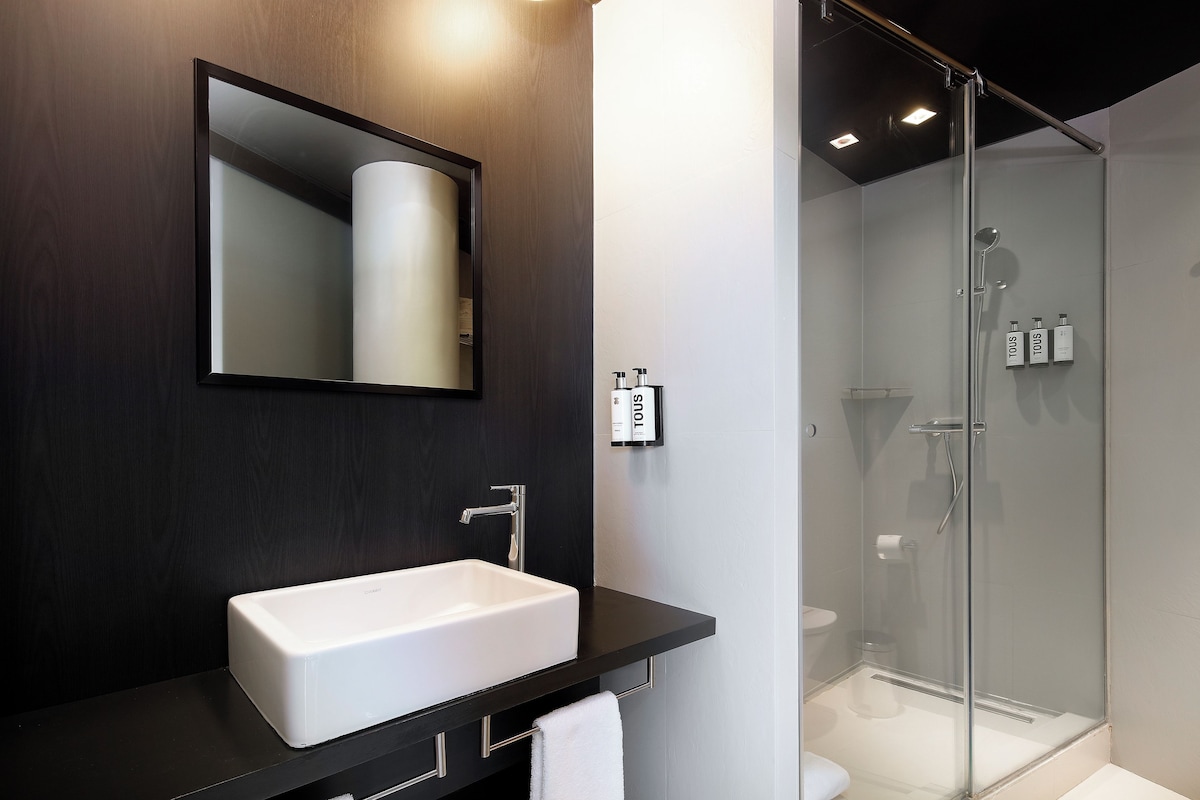 Un baño moderno con un lavamanos blanco, un espejo, cabina de ducha de vidrio y artículos de aseo en la pared.
