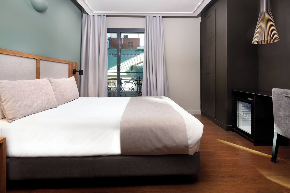 Une chambre d’hôtel moderne avec un grand lit, une petite télévision et un balcon donnant sur les bâtiments.