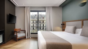 Quarto de hotel com uma cama grande, uma cadeira de madeira, uma televisão de tela plana e uma varanda com vista para edifícios.