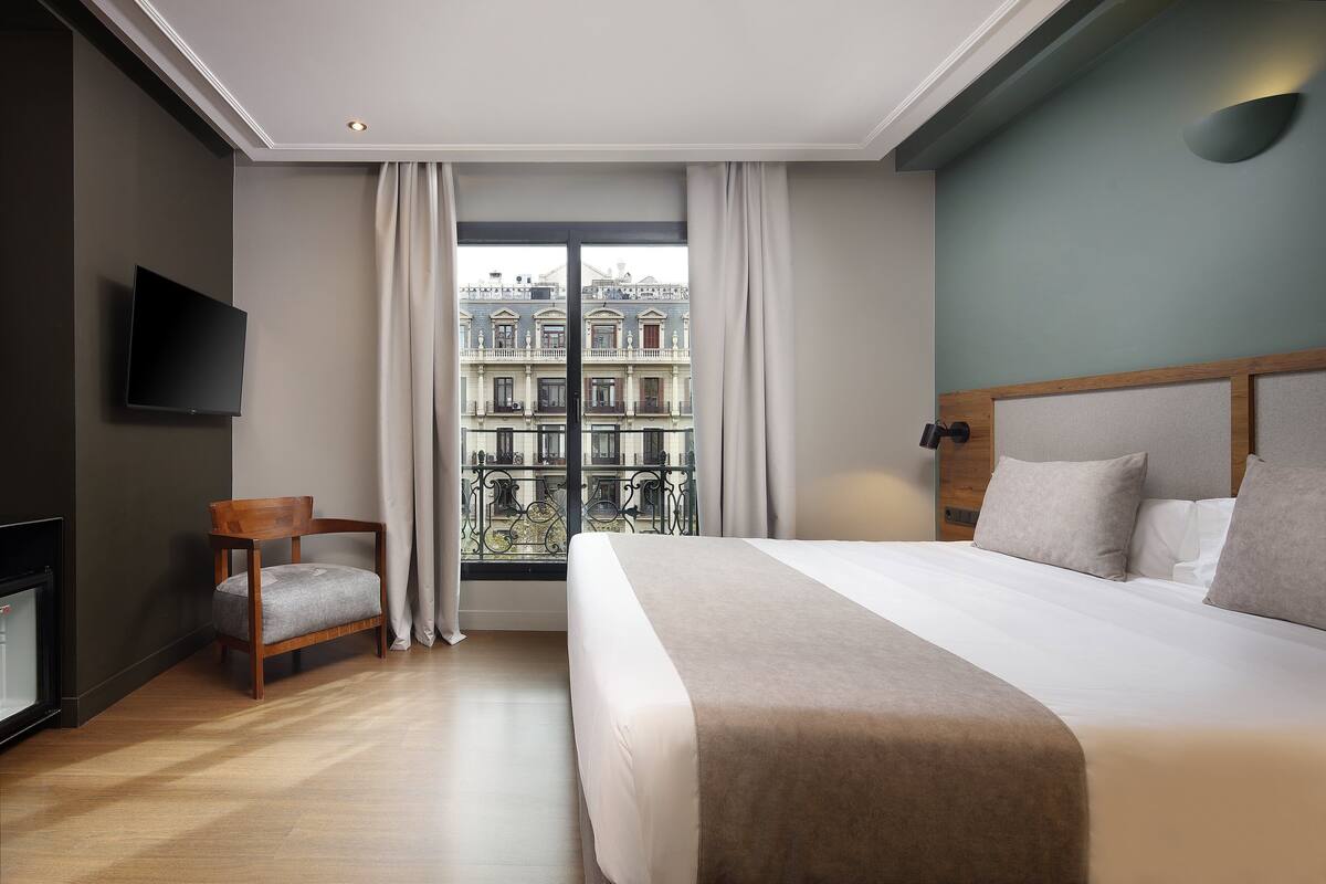 Une chambre d’hôtel avec un grand lit, une chaise en bois, un téléviseur à écran plat et un balcon offrant une vue sur les bâtiments.