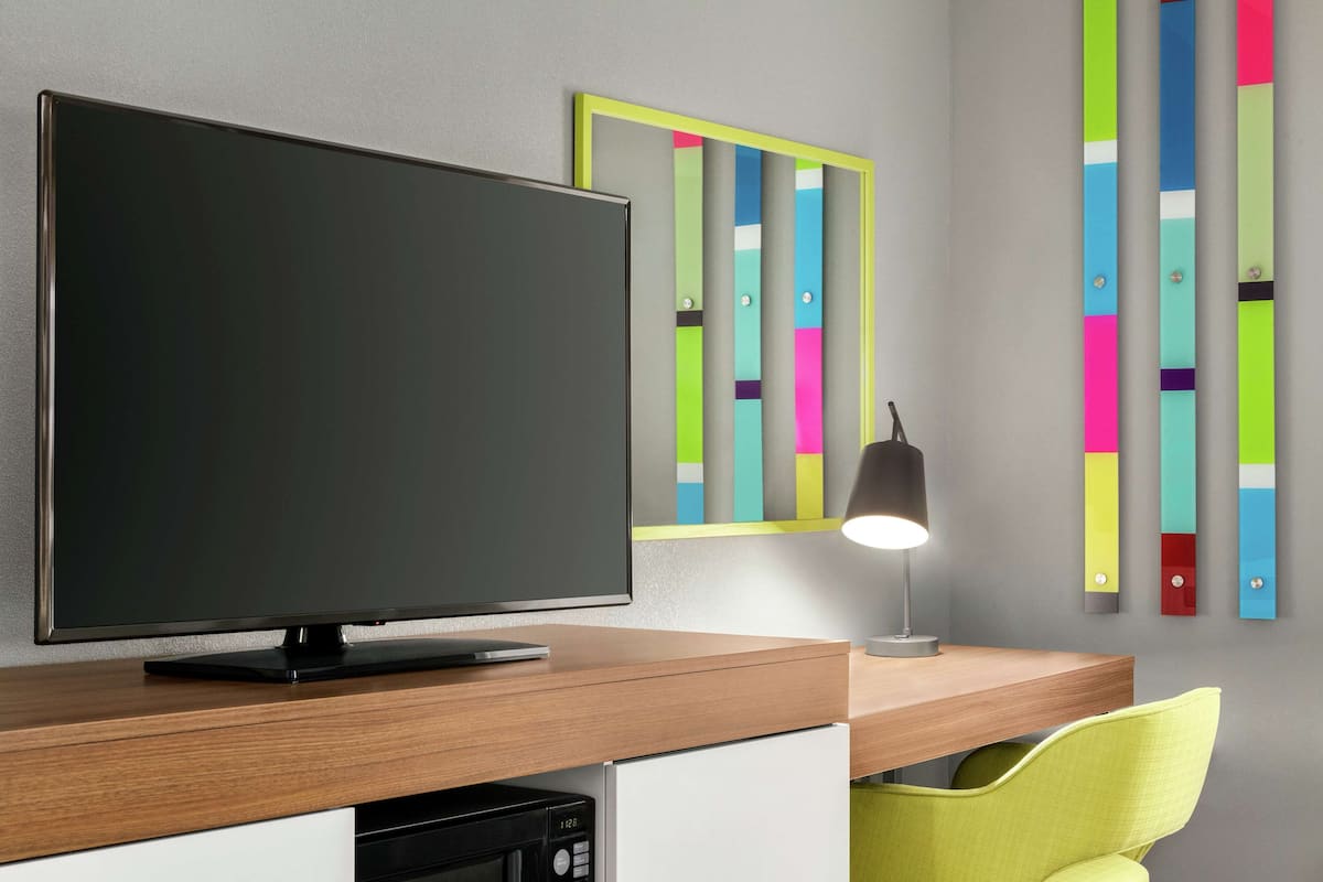 Una habitación moderna con una televisión de pantalla plana sobre un escritorio de madera, un refrigerador pequeño, una lámpara y un cuadro abstracto y colorido en la pared.