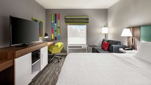 A hotel room with a flat-screen TV, a desk with a chair, a bed with a white comforter, a window with a striped blind, and a nightstand with a lamp. - Hampton Inn Oklahoma City-I-40 E. (Tinker Air Force Base) (Midwest City)