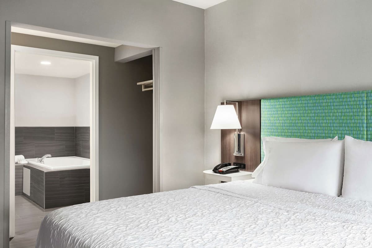 Habitación de hotel con cama, mesita de noche con lámpara, teléfono y baño con bañera y lavabo.