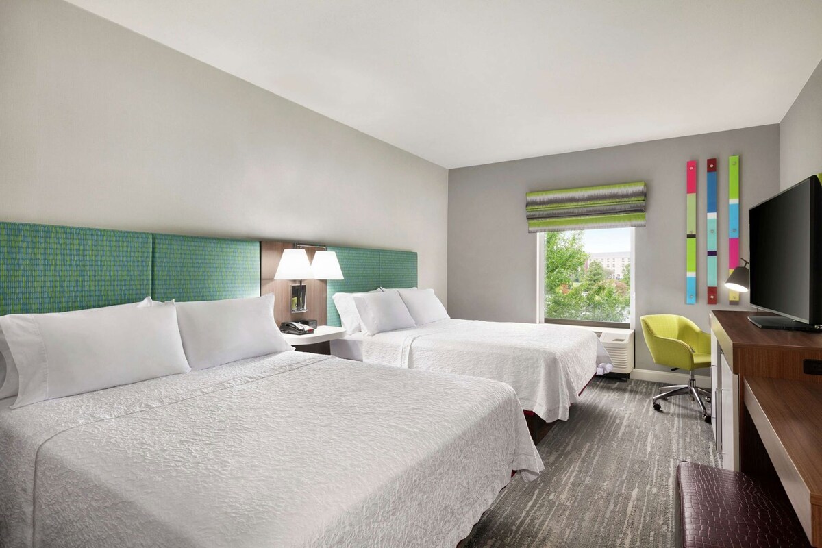 A hotel room with two beds, a desk, a chair, a TV, and a window with a view.