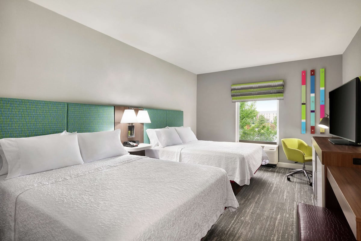 Habitación de hotel con dos camas, un escritorio, una silla, un televisor y una ventana con vista a árboles.
