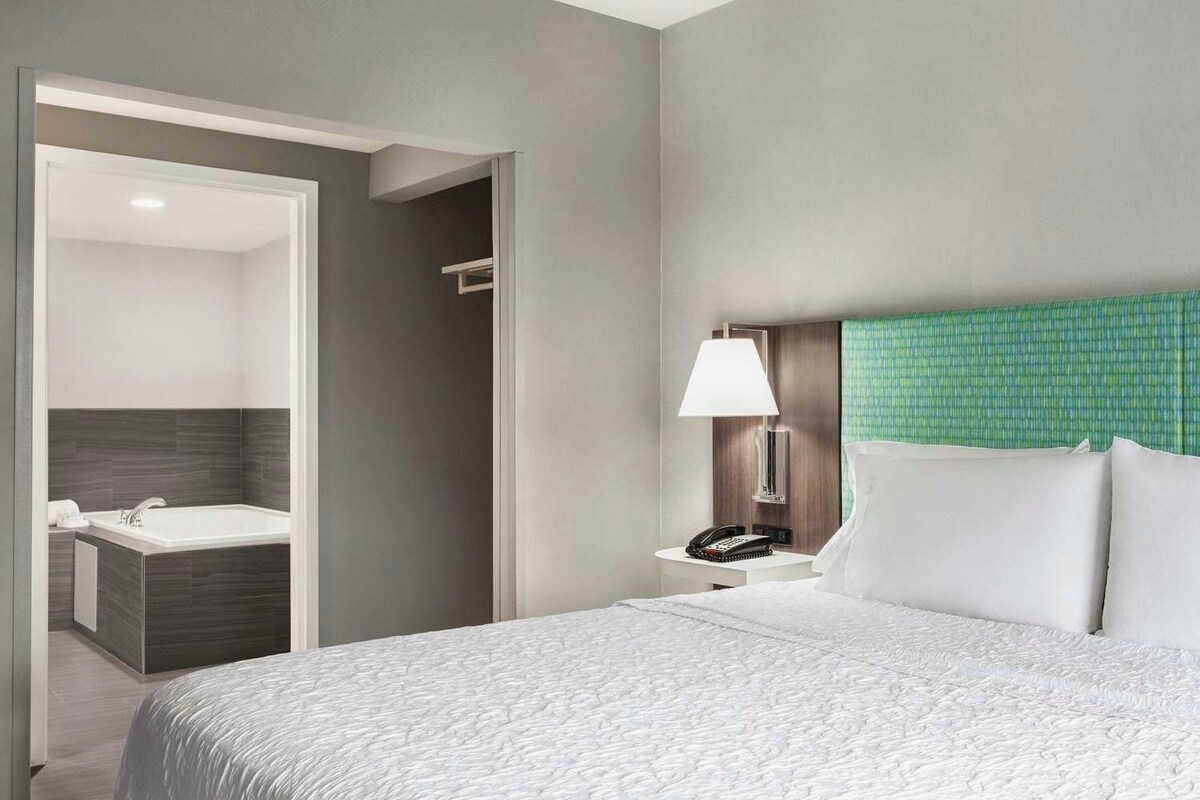 A hotel room with a bed, a nightstand, a lamp, a telephone, and a bathroom with a bathtub and a mirror.