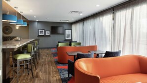 Ein modernes Hotelzimmer mit Holzboden, einer Bar mit Hockern, orangefarbenen Sitzgelegenheiten und einem großen Fenster mit Vorhängen.