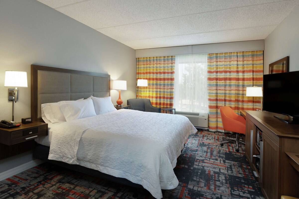 A hotel room with a large bed, a desk with a television, a chair, and a desk lamp.