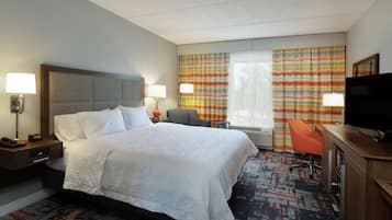 A hotel room with a large bed, a desk with a television, a chair, and a desk lamp.