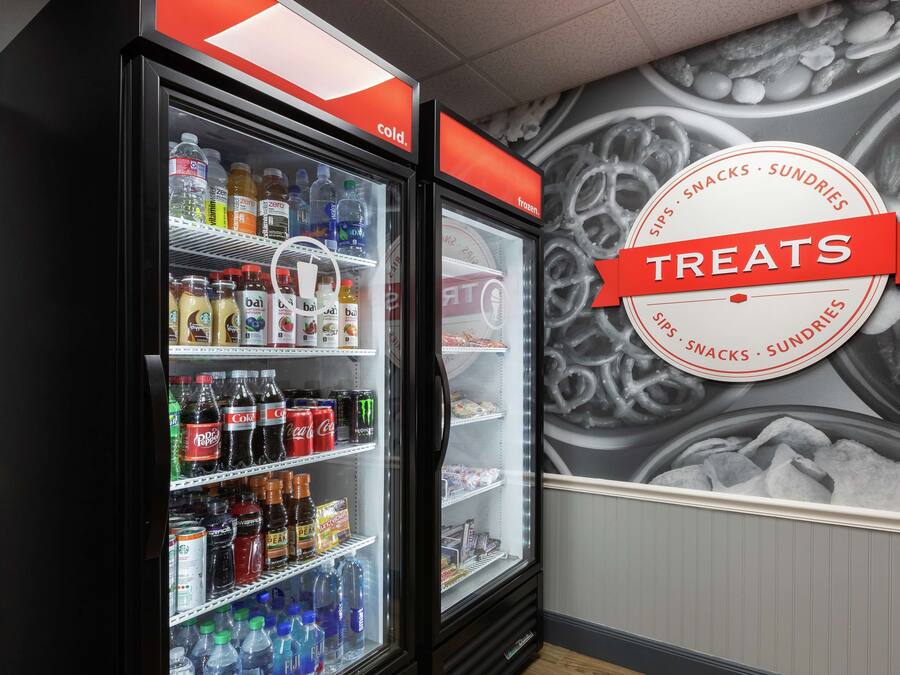 Vitrine refrigerada com diversas bebidas e lanches, e um letreiro na parede indicando 'TREATS' com um logotipo circular.