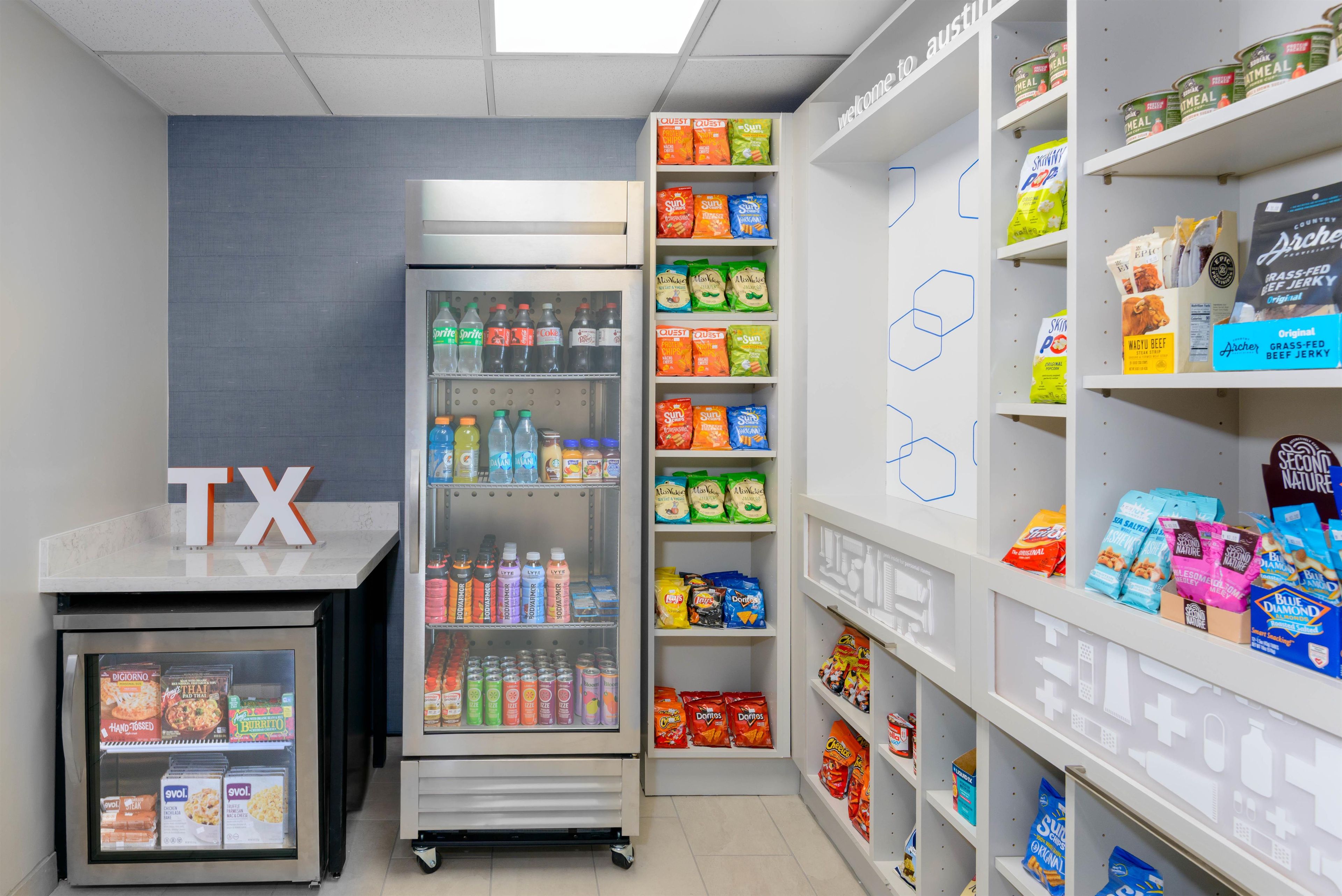 A self-service beverage and snack area with refrigerated drinks, a freezer, and shelves stocked with various snacks and beverages.