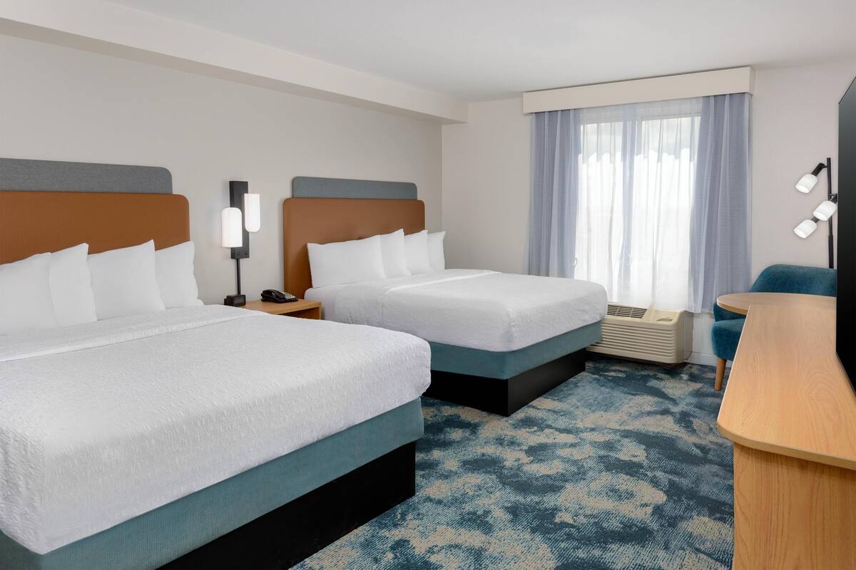 A hotel room with two beds, a desk, a chair, a lamp, and a window with curtains.