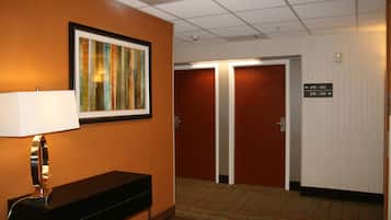 A hotel hallway with a framed artwork, a desk with a lamp, and double doors marked with room numbers.
