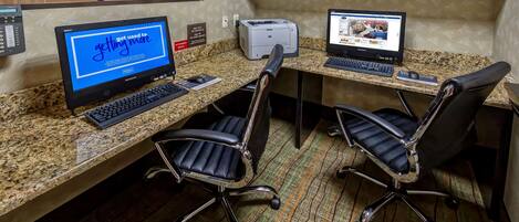 Un espace de travail dans un hôtel, comprenant deux bureaux, des ordinateurs, des chaises, une horloge et un tableau encadré accroché au mur.
