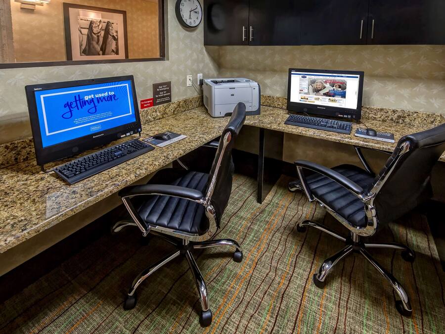 A hotel workspace with two desks, computers, chairs, a clock, and a framed picture on the wall.