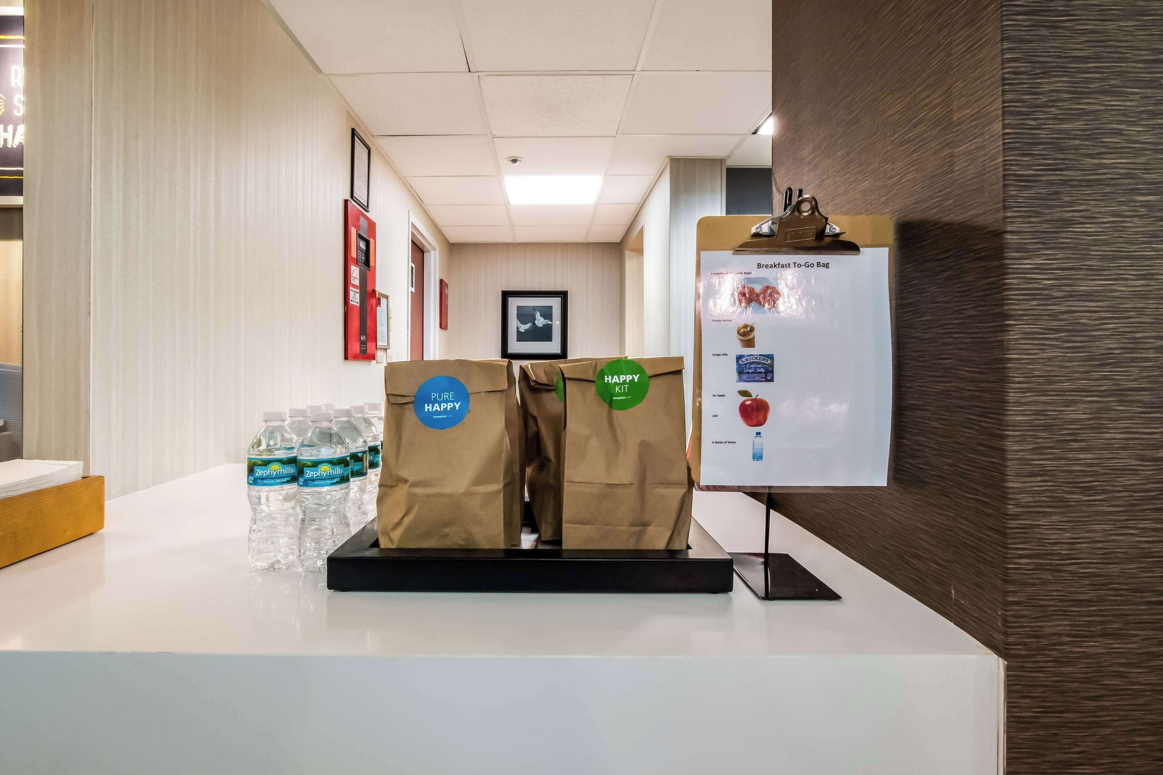 A reception area with a white counter, water bottles, a 'Happy Kit' display, and a 'Breakfast To-Go Bag' sign.