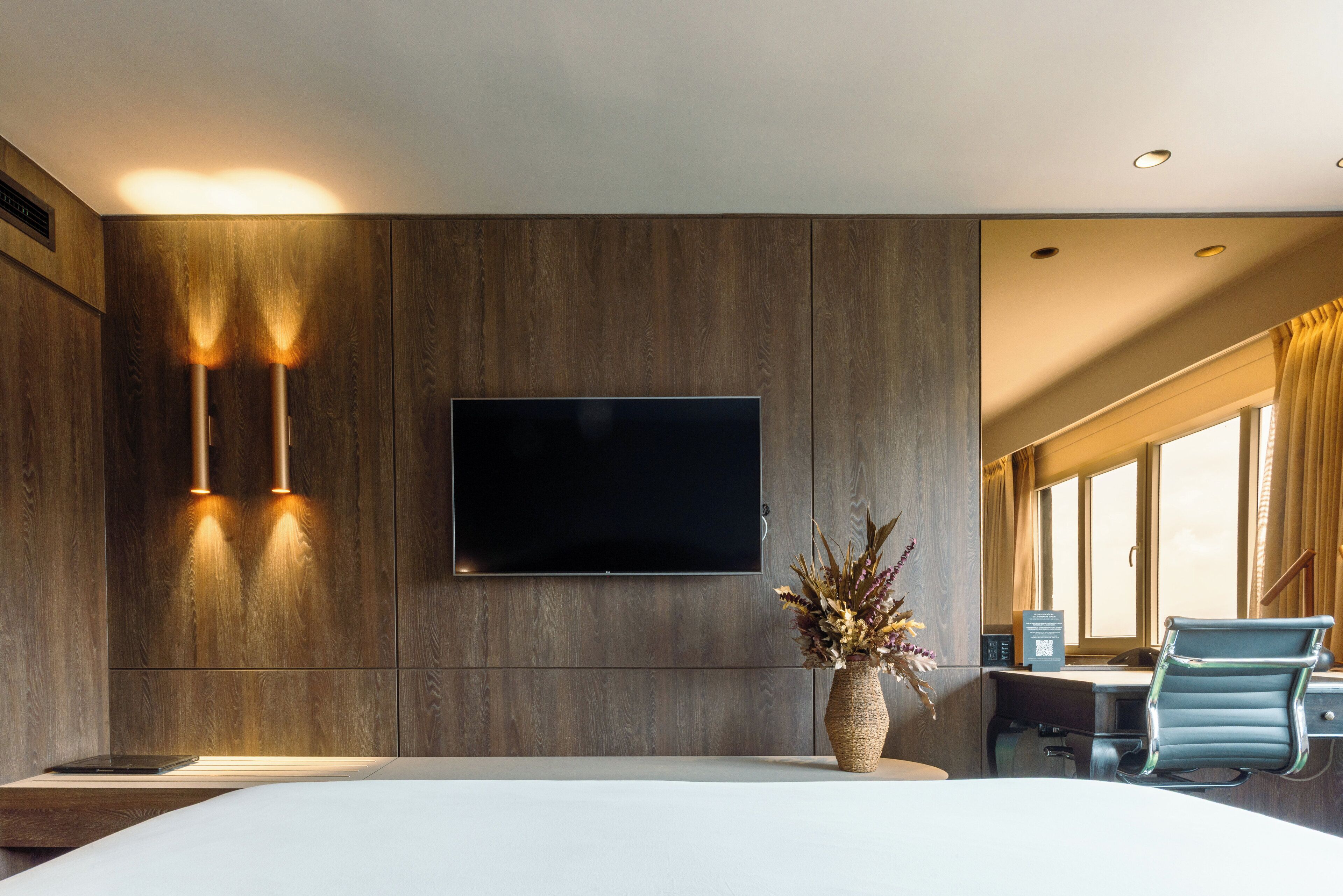 A modern hotel room with a large bed, a desk with a chair, a flat-screen TV mounted on the wall, and a vase of flowers on the desk.