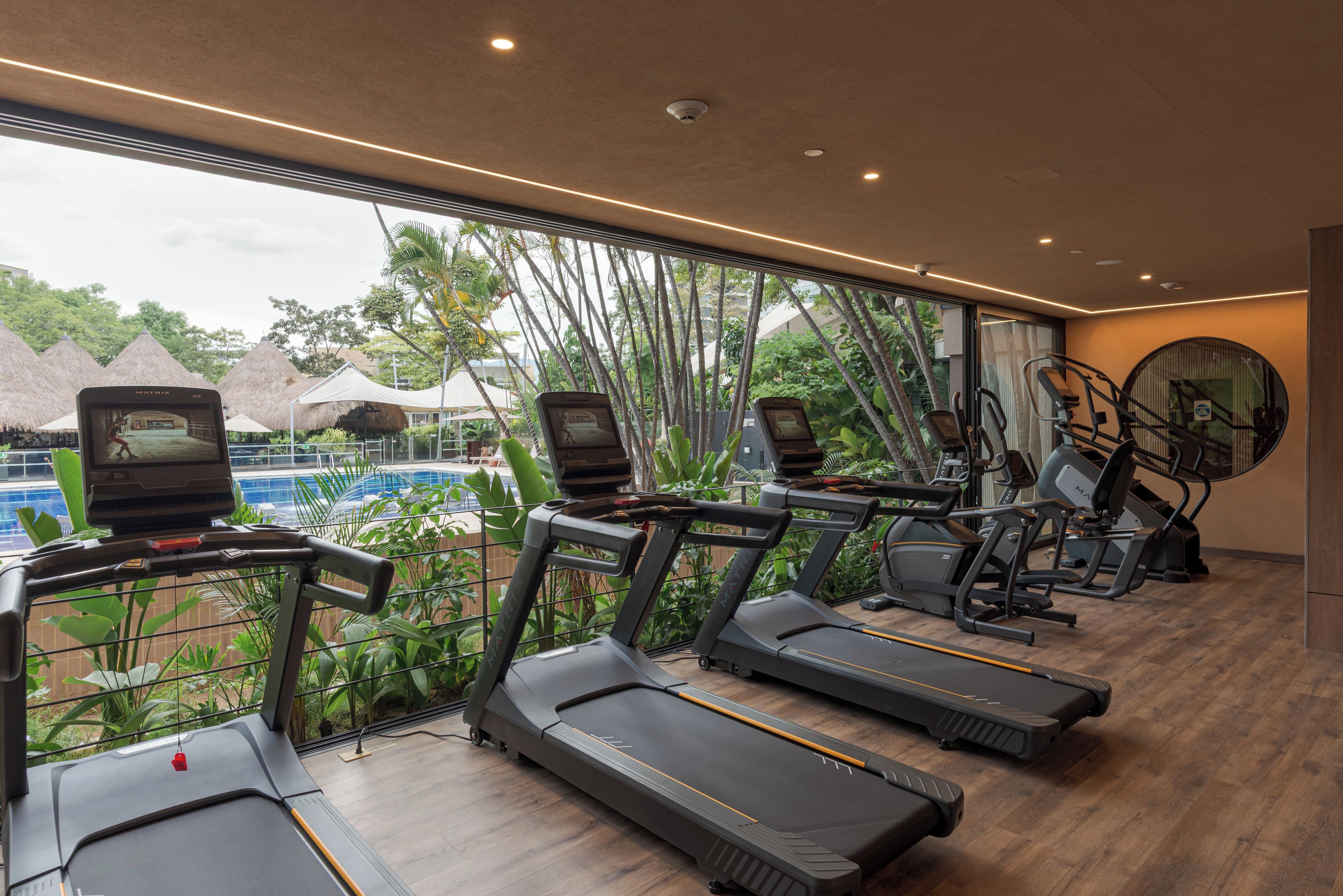 Academia com esteiras e equipamentos de ginástica, janelas amplas e vista para uma piscina e plantas tropicais.