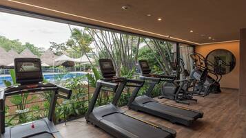 Academia com esteiras e equipamentos de ginástica, janelas amplas e vista para uma piscina e plantas tropicais.