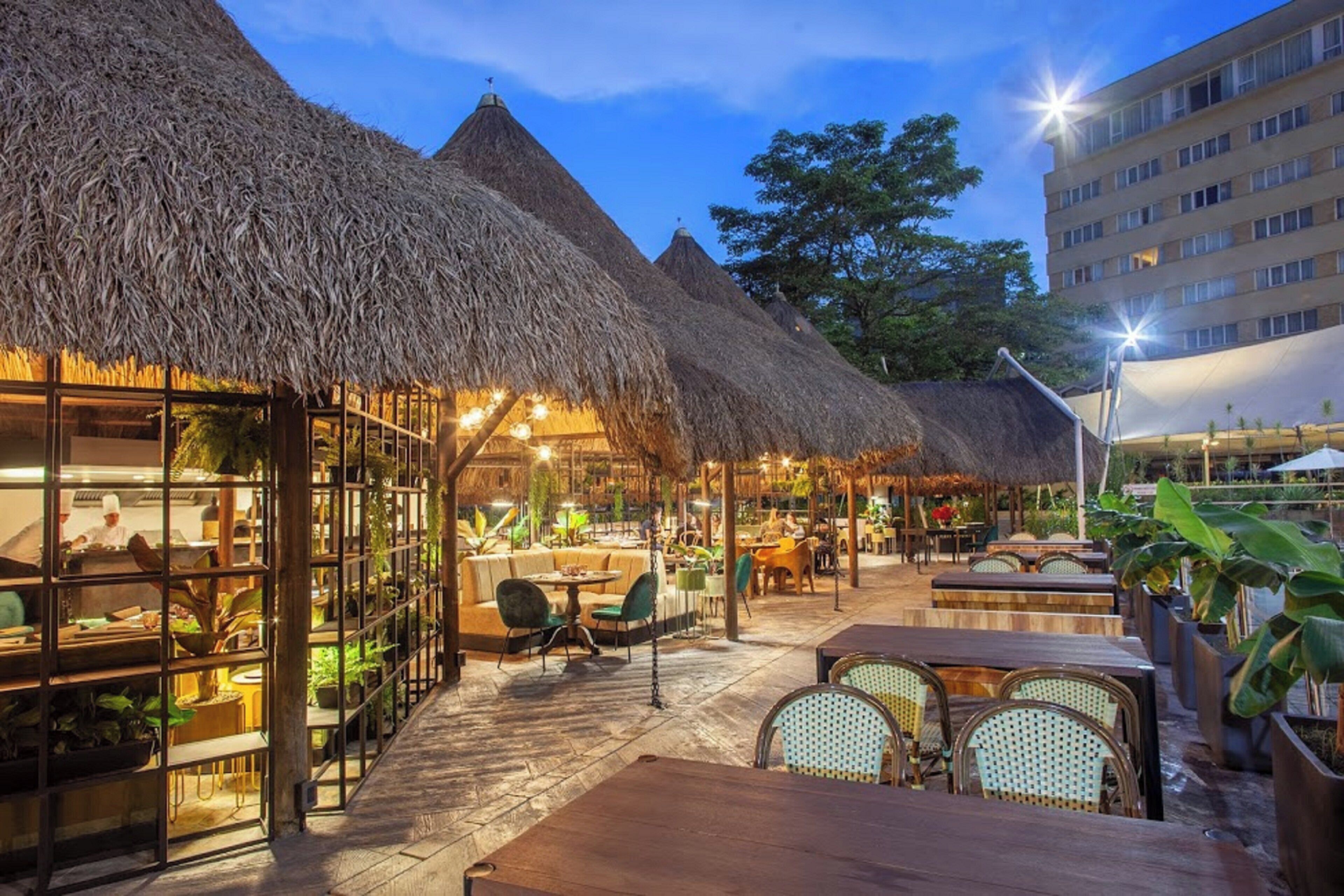 An outdoor restaurant with thatched roofs, tables, and chairs, surrounded by greenery and a building in the background.