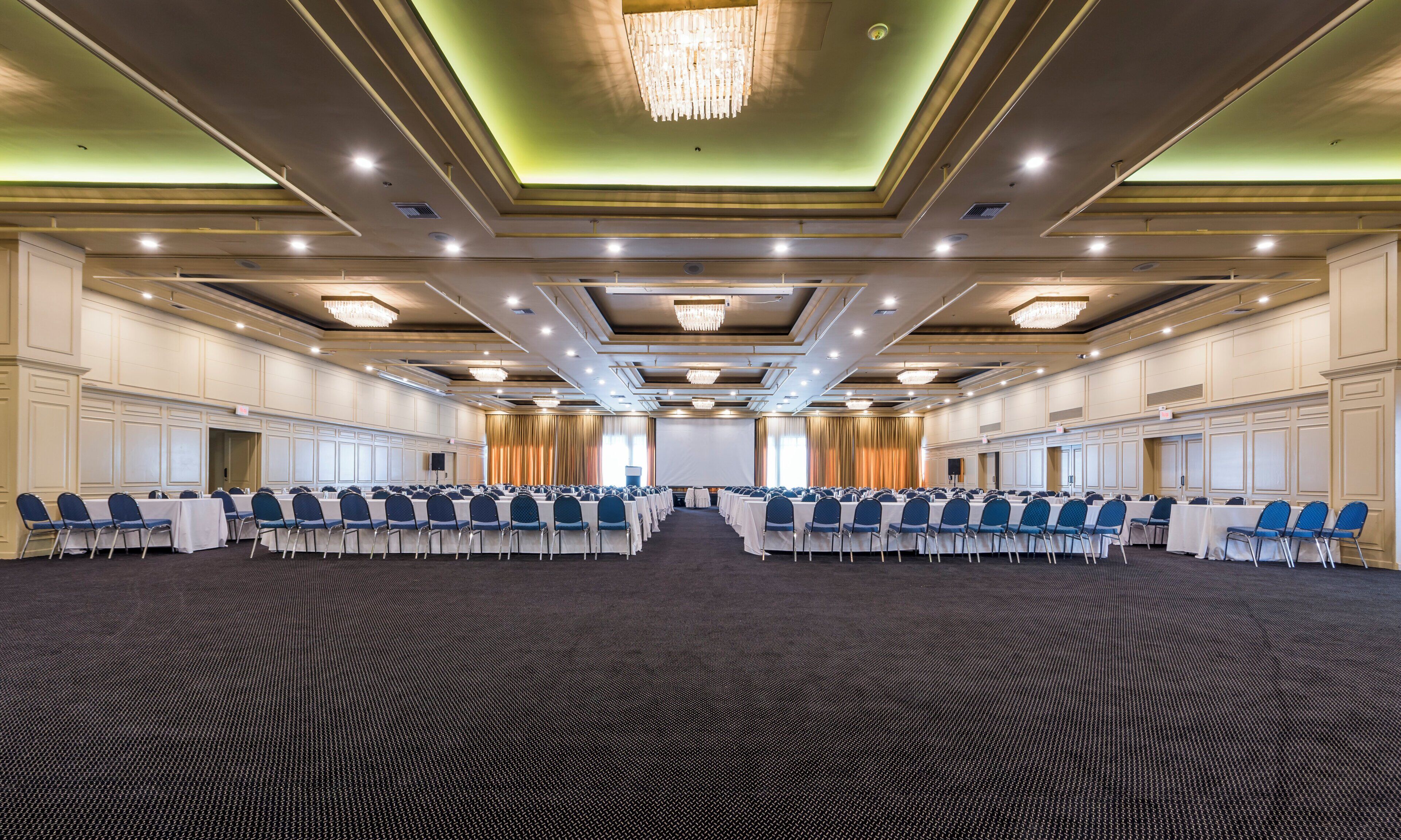 A large conference room with rows of tables and chairs, a stage with curtains, and a chandelier.