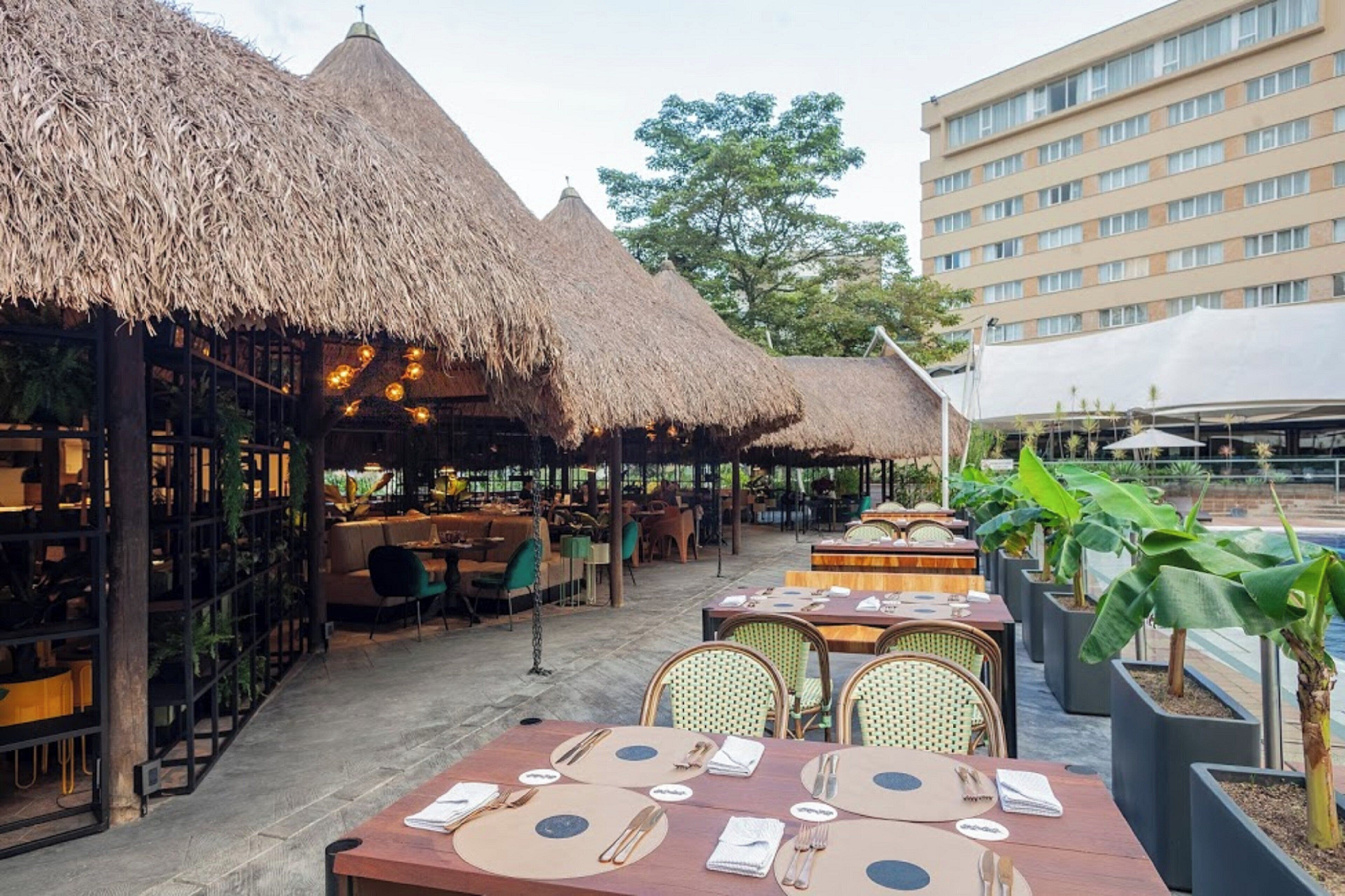 An outdoor restaurant area with thatched-roof structures, dining tables with set tables, and a modern building in the background.